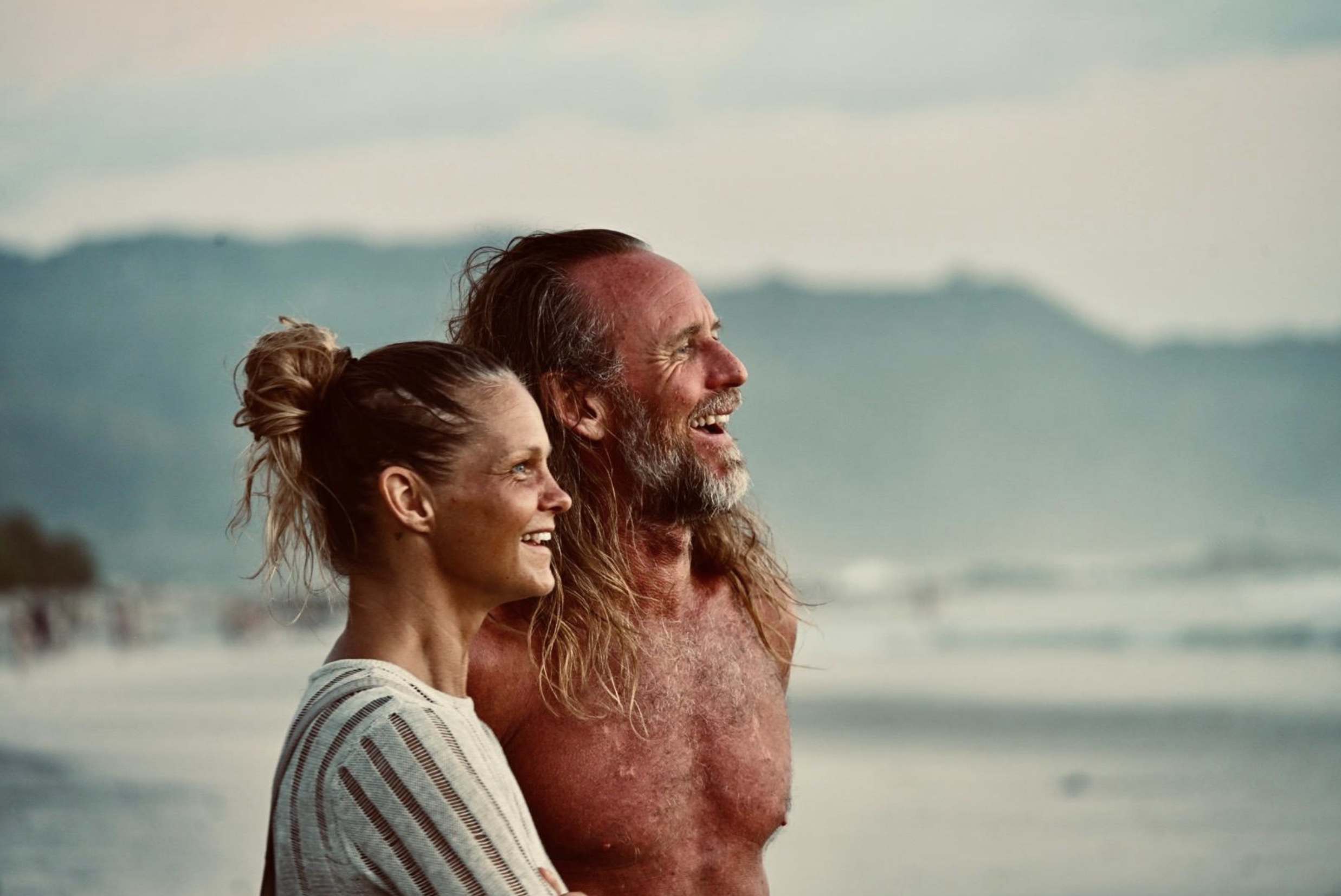 Christian og Carine smiler og ser utover havet på stranden, med tåkete fjell i bakgrunnen
